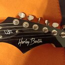 Černý hlavník elektrické kytary s šesti stříbrnými ladičkami. Na povrchu je bílý nápis „Harley Benton“ stylizovaným písmem a u matice logo „HB“. Povr…