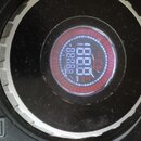 Detailní pohled na ovládací jog-wheel DJ kontroléru v černé barvě. Uprostřed je kulatý displej s červeným podsvícením a bílými digitálními číslicemi…