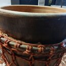 Detailní pohled na horní část dřevěného bubnu s tmavým povrchem. Nástroj je opleten tlustými oranžovými šňůrkami v pletené síťové struktuře. Na povrc…