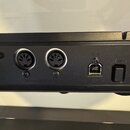Detailní pohled na zadní panel černého elektronického hudebního zařízení. Jsou vidět konektory s nápisy MIDI IN, MIDI OUT a USB, dále vstup POWER a D…