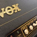 Detailní záběr na horní část kytarového komba s černým texturovaným povrchem. Na horní hraně je umístěno zlaté plastické logo VOX. Ovládací panel je…