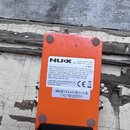 Zadní strana oranžového efekt pedálu značky NUX. Na bílém štítku je uveden web www.nuxefx.com, požadavek AC ADAPTER: DC 9V a text o shodě s pravidly…