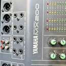 Detailní pohled na ovládací panel míchací konzole Yamaha MX200 v šedé barvě. Jsou vidět různé typy konektorů (XLR, jack, RCA) s popisky jako MONO OUT…