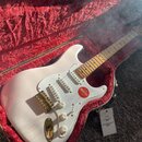 Bílá elektrická kytara typu Stratocaster s javorovým hnedým hmatníkem v otevřeném kufru s červenou plyšovou výstělou. Nástroj má bílý pickguard, tři…