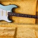 Světle modrá elektrická kytara typu Stratocaster s bílým pickguardem a hnědým hmatníkem. Na hlavě je viditelné logo Fender a nápis STRATOCASTER. Nást…