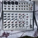 Stříbrný mixážní pult s černými ovládacími prvky. Na horním panelu je nápis EURORACK UB1202 a logo BEHRINGER. Zařízení obsahuje čtyři mikrofonní vstu…