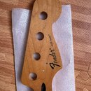 Dřevěný hриф basové kytary s přírodním povrchem a čtyřmi otvory pro ladicí strojky. Na hlavě je černý nápis Fender® a JAZZ BASS®. Povrch dřeva vykazu…