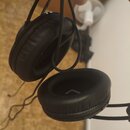 Černé on-ear sluchátka s polstrováním z umělé kůže. Jsou drátové a mají jednoduchý plastový rám. Na vnitřní straně jednoho z ušnic je vidět bílý symb…
