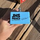 Světle modrý efekt pedál s nápisy JHS PEDALS a PAUL GILBERT PG-14. Vedle textu je umístěn černý podpis. Zařízení je v kovovém provedení s viditelným…