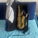 Otevřený modrý tvrdý kufýr s černým vnitřním polstrováním, ve kterém je umístěn zlatý saxofon. K nástroji patří bílá látková utěrka s modrým vzorem,…