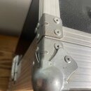 Detailní pohled na roh transportního kufru s hliníkovými lištami a černým povrchem. Roh je chráněn kovovoutýčovou rohovou výztuhou a spojovacími prvk…