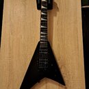 Černá elektrická kytara s tělem ve tvaru V umístěná na dřevěném stole. Na hlavě hmatníku je bílý nápis Jackson. Kytara má dva humbuckery, ladicí kolí…