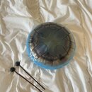 Modrý průhledný steelpan s černými vrypy tónových polí. Na povrchu je bílý nápis s označením tónů: G, F#, E, D, C, B, A. K nástroji jsou přiloženy dv…