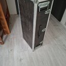 Černý transportní kufr s hliníkovými hranami a kovovými rohy. Má dvě madla, horní a boční, a tři kovové zámky. Povrch je viditelně opotřebovaný s něk…