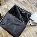 Otevřená černá textilní brašna s popruhem, položená na dřevěné podlaze. Vnitřní prostor je vyložen černým materiálem, na vnitřní straně víka se nachá…