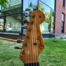 Detail hlavy elektrické kytary z dřeva s přirozeným povrchem. Na zadní straně je vidět logo Fender a nápis STRATOCASTER. Jsou zde šest ladicích kol v…