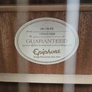 Detail vnitřní štítku akustické kytary přilepeného k dřevěnému korpusu. Na oválném štítku je uveden Model DR-100 EB a No. 17031312688. Je zde uveden…