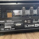 Zadní strana kytarového komba s černým povrchem a ochrannou mřížkou, za kterou jsou vidět elektronické součástky a elektrony. Na spodním černém panel…