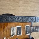 Detail elektrické kytary s vyznačenou dřevěnou strukturou a dvěma humbuckery v bílých rámečcích. Přes tělo kytary je položen černý kožený popruh s vy…