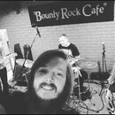 Černobílá fotografie zachycuje hudební kapelu v prostoru s cihlovou stěnou. V pozadí je vidět nápis Bounty Rock Cafe a tabule s textem Rockmax. Jsou…
