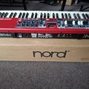 Červený digitální klavír s nápisem nord electro 6D leží na hnědé papírové krabici s logem nord®. Nástroj má bílé a černé klávesy, několik posuvníků,…
