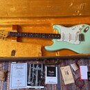 Světle zelená elektrická kytara typu Stratocaster s bílým pickguardem a javorovým hmatníkem v otevřeném hnědém kufru s žlutým polstrováním. Na hlavě…
