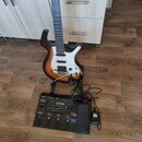 Elektrická kytara s trupem v barevně sunburst a bílým pickguardem stojí na stojanu. Před ní leží černý procesor s nápisy Roland GR-33 a GR Guitar Syn…