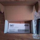 Otevřený hnědý karton, ve kterém leží bílý papírový manuál s nápisy SM57, INSTRUMENT MICROPHONE a logem SHURE. Vedle krabice leží černý dynamický mik…