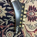 Detail hlavy elektrické kytary v černé barvě s viditelným logem Epiphone. Na hlavě jsou umístěny zlaté ladičky s bílými plastovými knoflíky. Povrch j…
