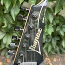 Detail hlavy elektrické kytary v černé barvě s bílým nápisem Ibanez a označením RG SERIES. Na hlavě jsou umístěny šest černých ladicích mechanismů. P…