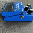 Modrý efekt pedál s nápisy Compression Sustainer CS-2, INPUT a OUTPUT. Na horní části je černý gumový spínač s logem BOSS. Pedál má tři ovládací knof…