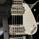 Detail elektrické kytary v černé barvě s bílým perleťovým pickguardem, na kterém je nápis GRETSCH. Na obou snímačích je vyryto U.S PAT 2892371. Vidit…