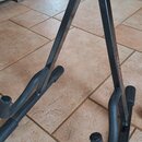 Černý kovový stojan na hudební nástroj v tvaru A s pěnovými ochrannými prvky. Na pravé noze je viditelný červený nápis "Hercules". Povrch stojanu vyk…