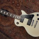 Elektrická kytara s bílým korpusem a hnědým hmatníkem. Na hlavě je viditelné logo Epiphone. Kytara má dva humbuckery, čtyři ovládací knoflíky a černý…