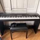 Černý digitální piano s klaviaturou a přívodním kabelem, umístěný na černém stojanu. Na levé straně je nápis MEDELI a vpravo označení PIANO 100. Před…