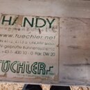 Detailní záběr na světlý dřevěný povrch s nalepenou papírovou nálepkou. Nálepka je zčásti odlepená a obsahuje zelený nápis „TUECHLER“ (otočený vzhůru…