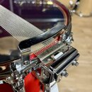 Detailní záběr na chromovaný snare strainer (napínák) u červeného bubnu. Na kovovém mechanismu je vyryt nápis "Pearl". Povrch chromu je lesklý a bez…