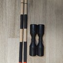 Dvě dřevěné bubenícké paličky s černými gumovými návleky a oranžovými prsteny u spodních černých konců. Vedle nich jsou dvě černé plastové nebo gumov…