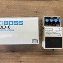 Fotografie zobrazuje efekt pedál Boss DD-6 Digital Delay v originální krabici. Pedál je stříbrný s černými nápisy Digital Delay, DD-6 a logem BOSS. N…