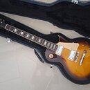 Elektrická kytara typu Les Paul v barvě sunburst s tmavě hnědým okrajem a žlutým středem, umístěná v černém tvrdém kufru. Na hlavě hmatníku je nápis…