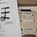 Fotografie ukazuje bílý pickguard s třemi instalovanými snímači a dvěma bílými oválnými knoflíky s nápisy VOLUME a TONE. Na snímačích je vidět nápis…