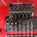 Detailní pohled na černý kovový kobylku s tremolo systémem na červeném povrchu kytary. Na horní části je vyryt nápis Licensed Under Floyd Rose Pats.…