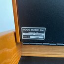 Detailní záběr na černý povrch hudebního nástroje s přilepeným bílým štítkem. Na štítku je uveden nápis MOOG MUSIC, Inc., ASHEVILLE, NC, minimoog® Vo…