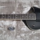 Černá elektrická kytara s tvarem připomínajícím Les Paul, umístěná v šedém polstrovaném kufru. Tělo má na pravé straně bílý grafický ornament a nápis…