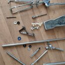 Sada kovových dílů a komponentů pro hudební stojany nebo držáky v stříbrné barvě. Na dřevěné podlaze jsou rozložené různé trubky, klouby, šrouby a je…