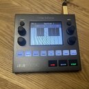 Šedý sampler s nápisem blackbox a logem 1010music v levém dolním rohu. Zařízení má uprostřed displej s názvem Section 1, čtyři otočné knoflíky a řadu…