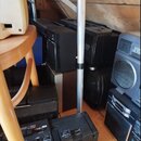 Fotografie zachycuje několik kusů starší audio techniky v černém plastovém provedení, umístěných v prostoru s dřevěnými stěnami. Na jednom z přístroj…