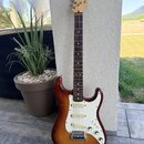 Elektrická kytara s trupem v červenohnědém sunburst provedení a bílým pickguardem. Na hlavě hmatníku je viditelné logo Fender a nápis STRATOCASTER. H…