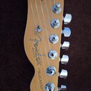 Detail hlavy elektrické kytary z přírodního dřeva. Na povrchu je černý nápis Fender, pod ním text MADE IN U.S.A. a níže nápis "TELECASTER". Jsou vidě…