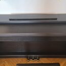 Černé digitální piano s dřevěnou texturou povrchu a přisluněným černým sedákem. Na horní desce i na spodní liště pod klávesnicí je vidět nápis KURZWE…